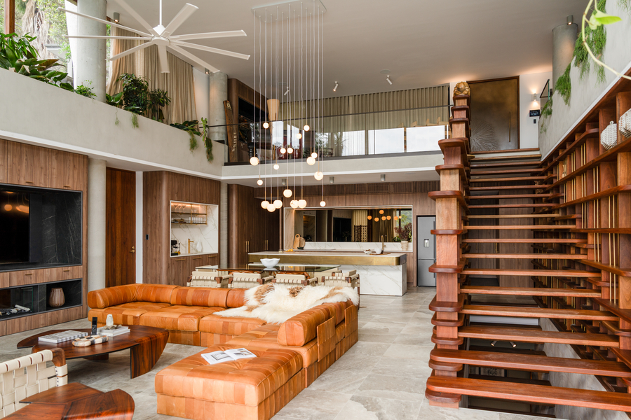 Stylish open-concept living room with a large leather sectional, modern pendant lighting, floating wooden staircase, and a sleek kitchen in the background.