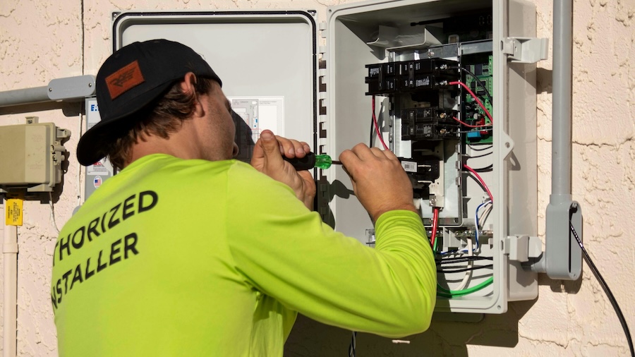 Electrician in a bright green shirt tightens a breaker in an open outdoor electrical panel during a whole-home upgrade.