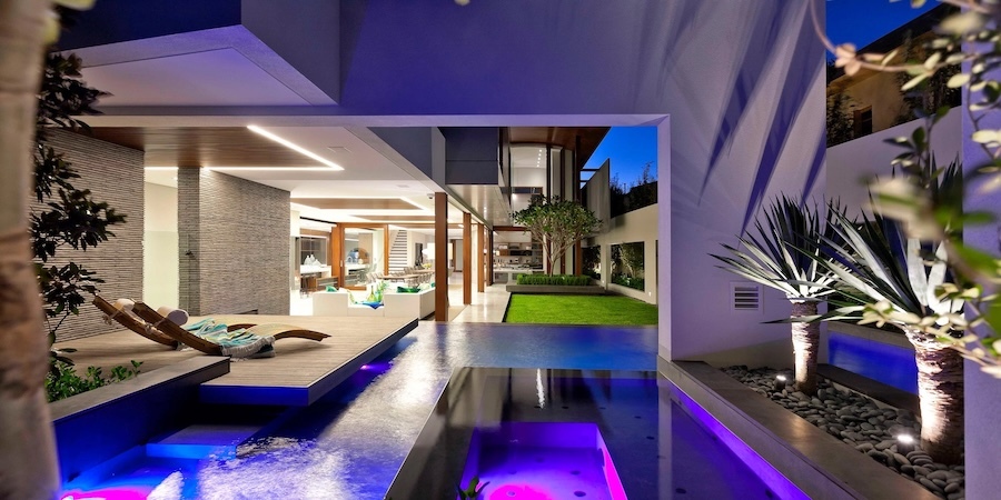 luxury home at dusk with illuminated pool, spa, wooden deck lounger, open living area, and courtyard garden.