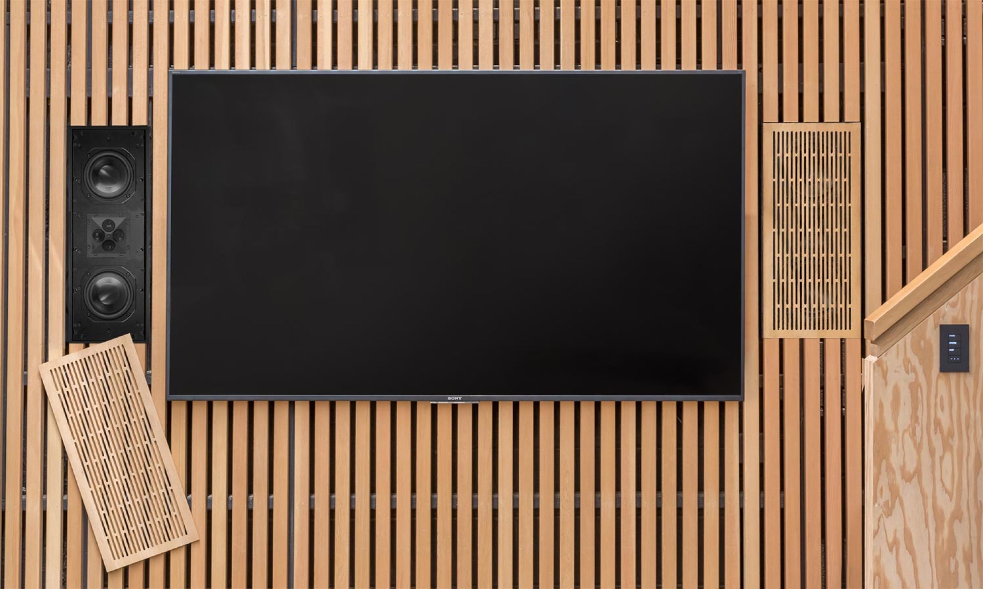 Hidden wall speaker with wooden slatted cover next to a large flat-screen TV