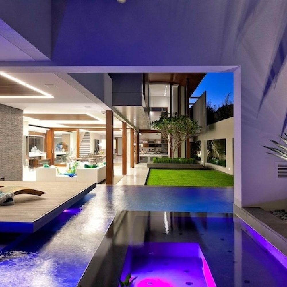 <span class="uk-h4">luxury home at dusk with illuminated pool, spa, wooden deck lounger, open living area, and courtyard garden.</span>