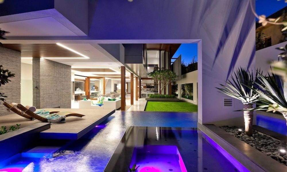 <span class="uk-h4">luxury home at dusk with illuminated pool, spa, wooden deck lounger, open living area, and courtyard garden.</span>