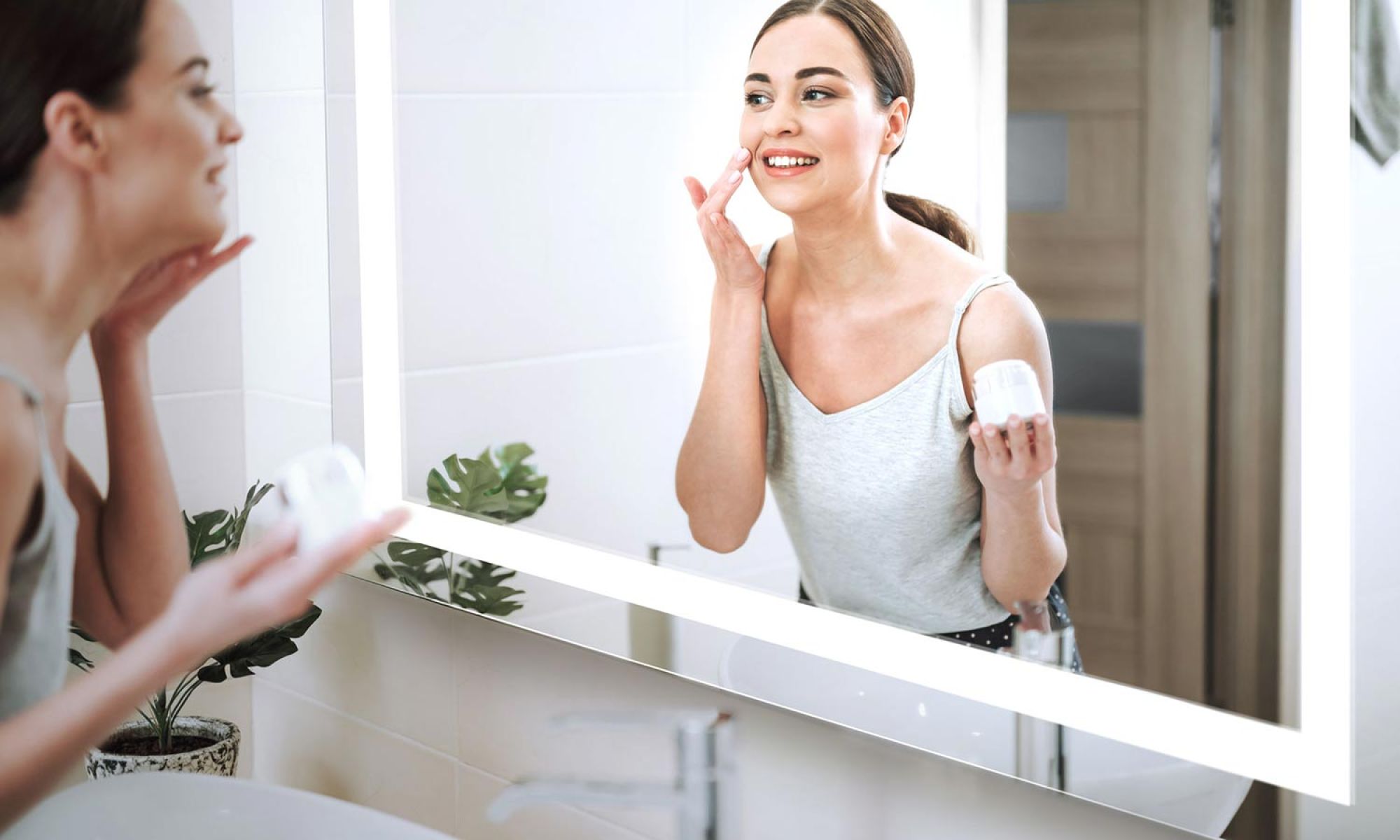 Woman smiling in front of a well-lit smart bathroom mirror while applying cream