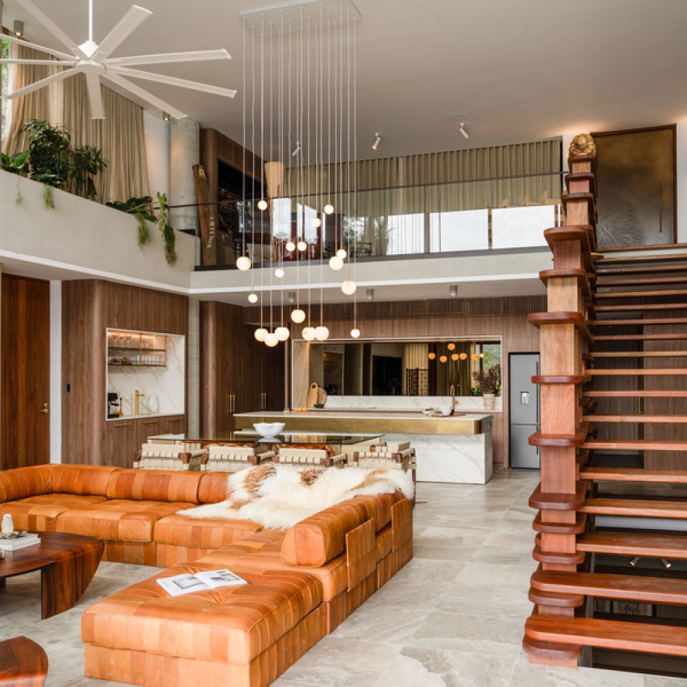 <span class="uk-h4">Stylish open-concept living room with a large leather sectional, modern pendant lighting, floating wooden staircase, and a sleek kitchen in the background.</span>